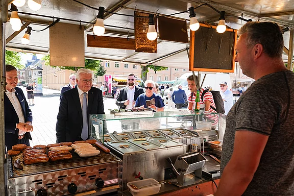 Ein Grillstand auf einem Markt, an dem verschiedene Würstchen zubereitet werden. Ein Verkäufer steht hinter dem Stand und spricht mit Gästen, die sich die Angebote ansehen. Im Hintergrund sind mehrere Personen, die interessiert zuschauen.