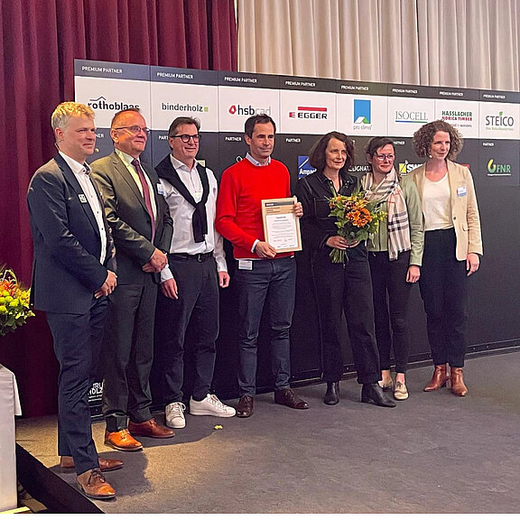 Eine Gruppe von sechs Personen steht auf einer Bühne vor einem Banner mit verschiedenen Logos. In der Mitte hält ein Mann ein Zertifikat, umgeben von anderen Personen, die Blumen und Urkunden halten. Die Atmosphäre wirkt feierlich und stolz.