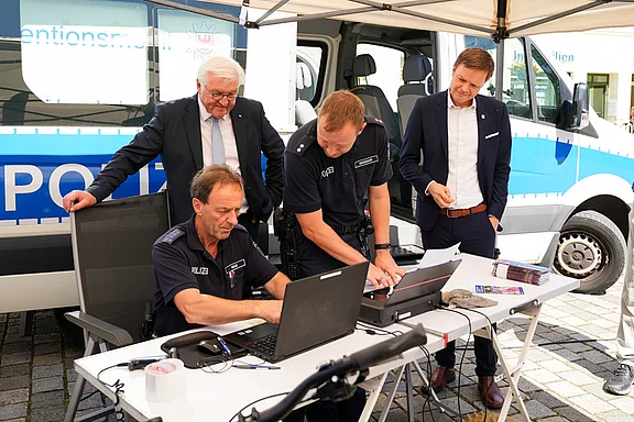 Zwei Polizisten arbeiten an einem Laptop, während zwei weitere Personen, darunter ein Mann in einem Anzug, die Situation beobachten. Im Hintergrund steht ein Polizeifahrzeug. Die Szene zeigt eine Polizeiveranstaltung zur Aufklärung oder Information.