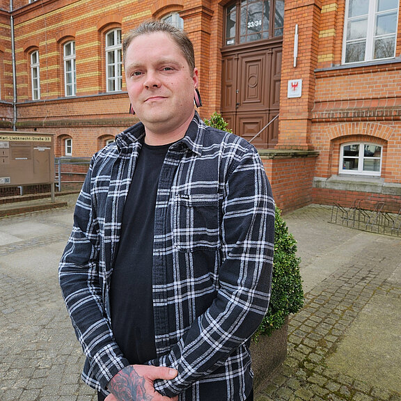Ein Mann mit kurzen, aufgestellten Haaren und Ohrsteckern steht vor einem historischen Gebäude mit Backsteinfassade. Er trägt ein karierte Jacke über einem schwarzen T-Shirt und hat bunte Tattoos an den Unterarmen. Im Hintergrund ist eine Pflanze und der Eingang des Gebäudes zu sehen.