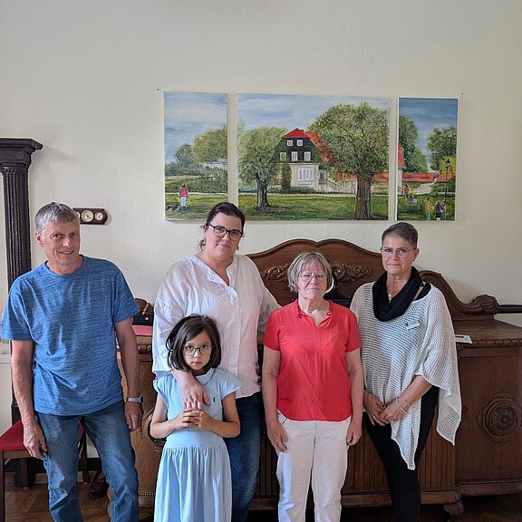 Gruppenfoto in Innenraum: fünf Personen stehen vor einer dreiteiligen Landschaftsmalerei mit Haus und Bäumen. Hinten vier Erwachsene, vorn ein Mädchen im blauen Kleid, die Arme verschränkt. Stimmung neutral.