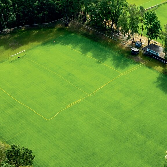 Luftaufnahme eines leeren Fußballplatzes mit sattem Grün. Gelbe Bewässerungsschläuche liegen in Rechtecken über dem Rasen. Ein Tor ist sichtbar. Am Rand säumen Bäume und wenige kleine Gebäude den Platz.