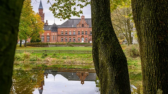 Ein historisches, rotes Gebäude mit Türmen und Fenstern spiegelt sich im Wasser eines Teiches. Umgeben von grüner Landschaft und Bäumen, vermittelt die Szene eine ruhige, malerische Atmosphäre.