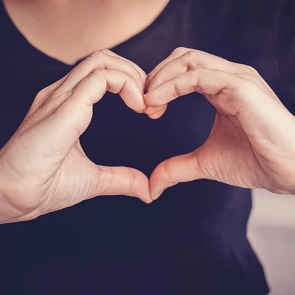 Zwei Hände formen ein Herzsymbol, das die Darstellung von Liebe und Zuneigung vermittelt. Der Hintergrund ist unscharf, sodass der Fokus auf den Händen liegt.