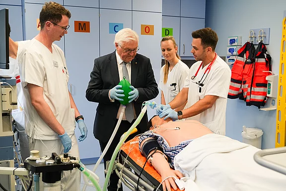 Eine Gruppe von vier Personen in einem Krankenhauszimmer. Ein älterer Mann in einem Anzug hält ein Beatmungsgerät, während Ärzte und Pflegekräfte an einem Patienten arbeiten, der auf einer Liege liegt. Alle sind auf die medizinische Versorgung des Patienten konzentriert.