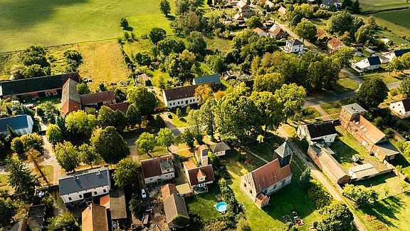 Vogelperspektive auf ein ländliches Dorf mit mehreren Häusern, umgeben von Bäumen und Wiesen. Ein kleiner Pool ist sichtbar. Im Hintergrund sind weitere Häuser und eine Kirche zu erkennen, die in eine grüne Landschaft eingebettet sind.