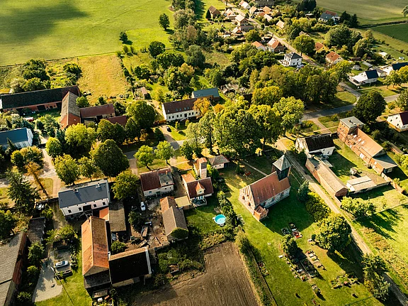 Vogelperspektive auf ein ländliches Dorf mit zahlreichen Häusern, umgeben von grünen Feldern und Bäumen. In der Mitte ist eine Kirche mit einem spitzen Turm sichtbar. Einige Gärten sind mit Pflanzen ausgestattet, und ein Pool ist im Garten eines Hauses zu sehen.