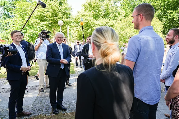 Eine Gruppe von Menschen steht im Freien, während ein älterer Mann mit Brille, der freundlich lächelt, im Mittelpunkt steht. Um ihn herum stehen mehrere Personen, die ihn anschauen und zuhören. Kamerateams sind ebenfalls anwesend und nehmen die Szene auf.