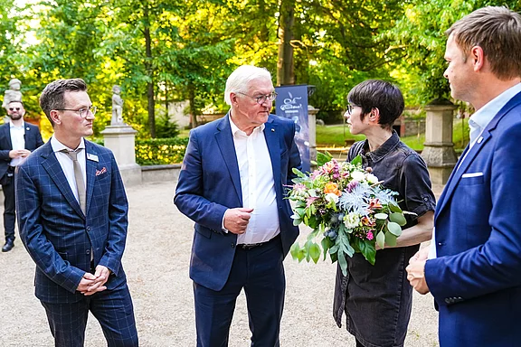 Vier Personen stehen im Freien, umgeben von Bäumen. Eine Person hält einen Blumenstrauß, während die anderen sie anlächeln und aufmerksam zuhören. Die Atmosphäre wirkt freundlich und festlich.