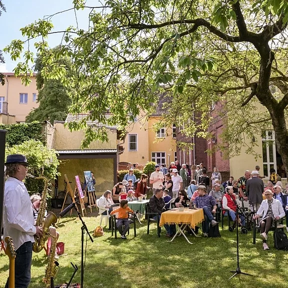 Gartenfest mit Live-Musik: Viele Menschen sitzen an Tischen unter einem großen Baum, unterhalten sich und hören einem Saxofonisten zu. Mikrofone und Instrumente stehen bereit. Im Hintergrund Wohnhäuser; freundliche, sonnige Atmosphäre eines nachbarschaftlichen Kultur- oder Sommerfestes.