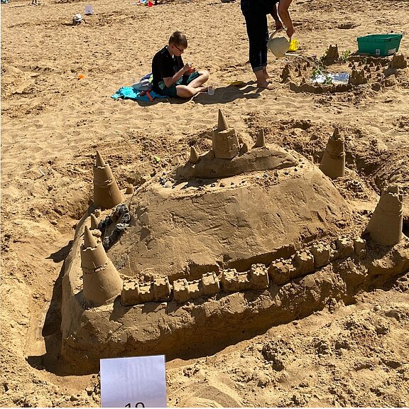 Große Sandburg am Strand: ein hoher Hügel mit mehreren spitzen Türmen, verbunden durch eine umlaufende Mauer und kleine Zinnen. Kinder arbeiten im Hintergrund weiter im Sand. Sonniger Tag.
