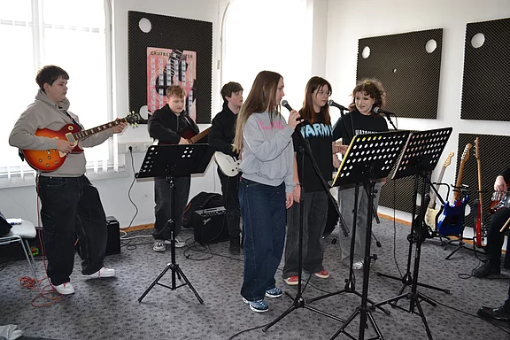 Eine Jugendband probt in einem Raum: Zwei singen, mehrere spielen E‑Gitarre und Bass vor Notenständern, Verstärkern und Mikrofonständern. Atmosphäre einer konzentrierten Bandprobe.