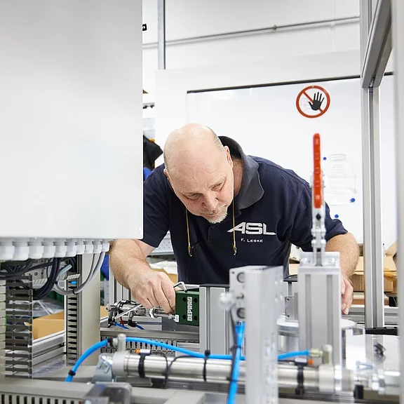 Techniker in einer Werkhalle richtet eine industrielle Maschine ein. Er beugt sich über das offene Gerät, prüft Bauteile und Kabel und justiert ein Messmodul. Helle, saubere Umgebung, Fokus auf präziser Montagearbeit.