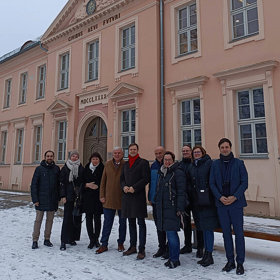 Eine Gruppe von zehn Personen steht vor einem historischen, hellrosa Gebäude im Schnee. Einige tragen Winterkleidung, während andere Schals und Mäntel tragen. Die Gruppe lächelt und posiert freundlich für das Foto.