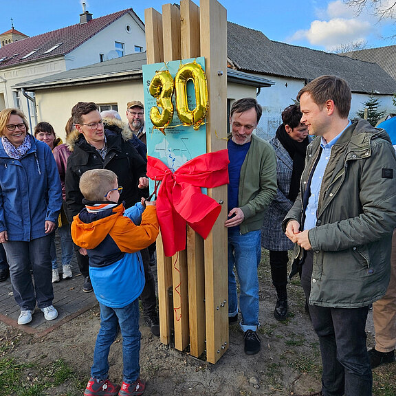 Eine Gruppe von Menschen steht zusammen, während ein Junge mit einer roten Schleife ein Holzschild mit der Zahl „30“ anfasst. Die Umgebung zeigt eine Feierlichkeit, und die Anwesenden lächeln und schauen gespannt auf das Schild.