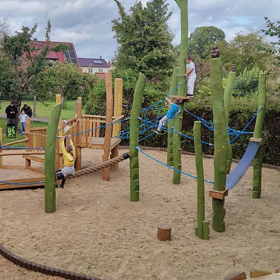Ein Spielplatz mit Holzkonstruktionen in der Mitte, umgeben von Rasen und Bäumen. Kinder spielen auf verschiedenen Geräten: einige klettern auf die Pfosten, andere schwingen an Seilen oder balancieren auf einem Brett. Sandboden sorgt für sichere Landung. Wolkiger Himmel im Hintergrund.