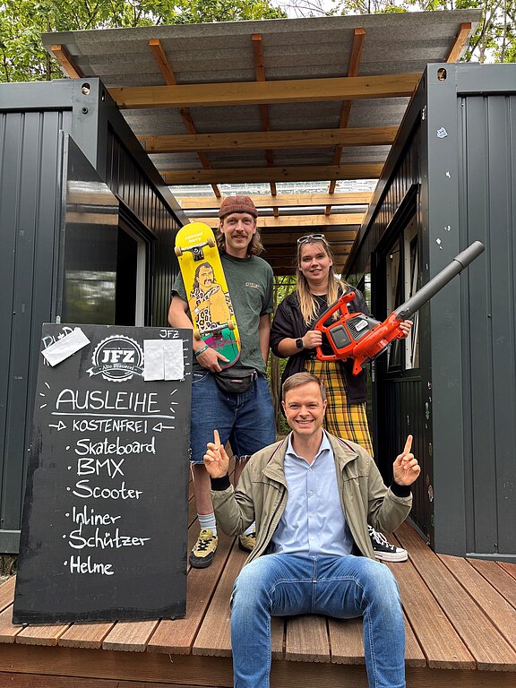 Drei Personen posieren vor einem Containereingang neben einer Tafel mit kostenloser Ausleihe: Skateboard, BMX, Scooter, Inliner, Schützer, Helme. Eine Person hält ein Skateboard; vorne sitzt jemand und zeigt nach oben.