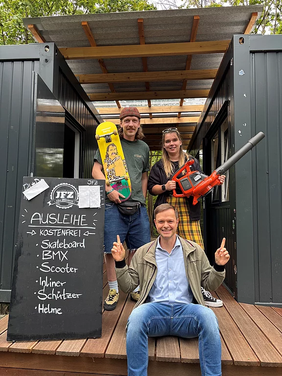 Drei Personen posieren vor einem Containereingang neben einer Tafel mit kostenloser Ausleihe: Skateboard, BMX, Scooter, Inliner, Schützer, Helme. Eine Person hält ein Skateboard; vorne sitzt jemand und zeigt nach oben.