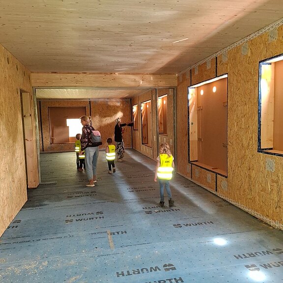 Kinder in leuchtend gelben Warnwesten gehen mit zwei Erwachsenen durch einen Rohbau aus Holz. Offene Tür- und Fensteröffnungen, unvollständige Wände und Schutzfolie am Boden zeigen die Baustelle. Die Gruppe besichtigt sicher geführt den Innenraum.