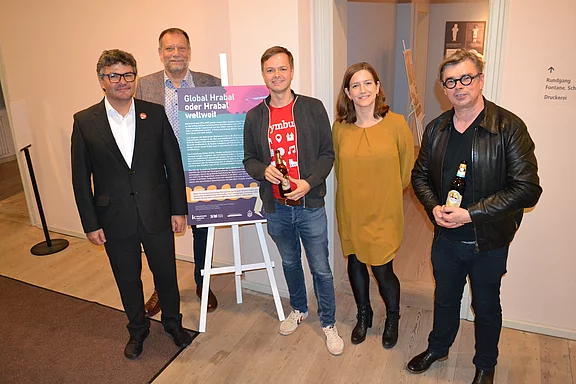 Eine Gruppe von fünf Personen steht vor einem Informationsplakat mit dem Titel „Global Hrabal oder Hrabal weltweit“. Sie lächeln und halten Getränke. Der Raum hat schlichte Wände und einen hellen Boden, Hintergrundelemente sind minimal.