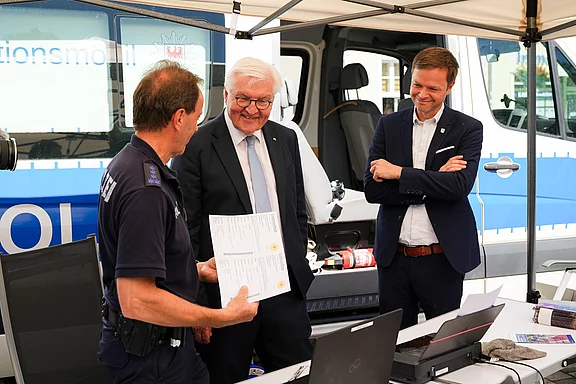 Der Bundespräsident steht in einem Gespräch mit einem Polizisten, der Unterlagen zeigt. Ein weiterer Mann beobachtet das Geschehen mit Interesse. Im Hintergrund ist ein Polizeifahrzeug sichtbar. Die Szene vermittelt einen Austausch über Sicherheit oder öffentliche Dienste.