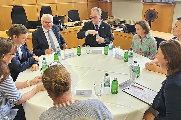 Eine Gruppe von acht Personen sitzt an einem runden Tisch in einem Besprechungsraum. Sie diskutieren bzw. hören einem Redner zu. Auf dem Tisch stehen Wasserflaschen und einige Papiere. Der Raum hat große Fenster und eine Holzwand im Hintergrund.