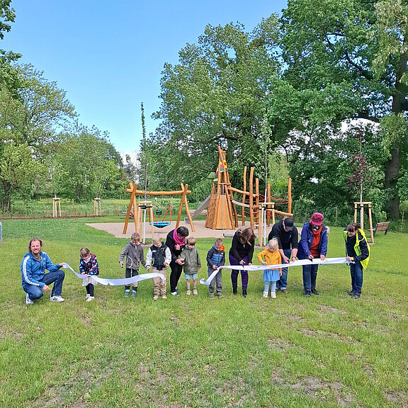 Eine Gruppe von Kindern und Erwachsenen hält gemeinsam ein langes Banner auf einer Wiese in einem Park. Im Hintergrund sind Spielgeräte und Bäume zu sehen. Die Szene vermittelt ein Gefühl von Gemeinschaft und Freude.