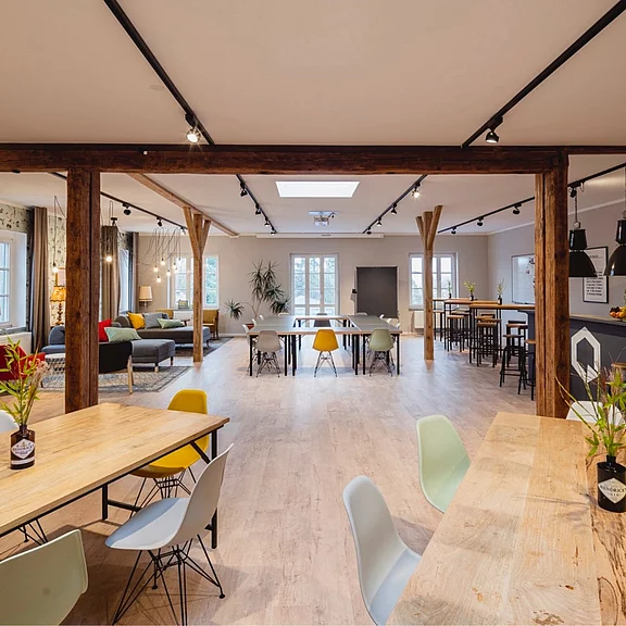 Heller, großzügiger Raum mit Holzboden und sichtbaren Balken: mehrere Tische mit bunten Stühlen, hohe Stehtische, Lounge mit Sofas und Pflanzen. Große Fenster und Deckenlichter sorgen für viel Tageslicht; geeignet für Treffen, Arbeiten oder Veranstaltungen.