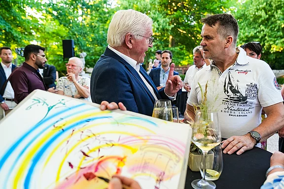 Zwei Männer führen ein Gespräch bei einer Veranstaltung im Freien. Im Hintergrund sind weitere Personen und eine grüne Umgebung zu sehen. Ein bemaltes Blatt Papier mit bunten Farben wird im Vordergrund gehalten. Getränke stehen auf einem Tisch.