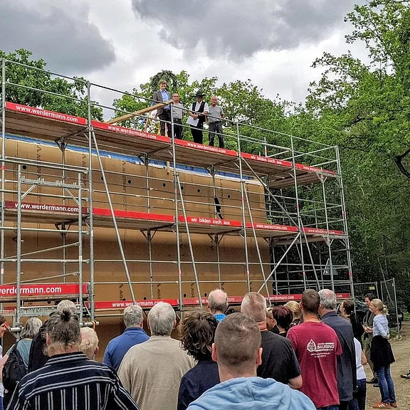 Vor einem großen Gerüst versammelt sich eine Gruppe von Menschen. Oben auf dem Gerüst sprechen drei Personen zur Menge – eine feierliche Baustellenzeremonie, vermutlich Richtfest, unter freiem Himmel.