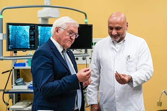 Ein Mann in einem Anzug und ein Arzt in einem weißen Kittel stehen in einem medizinischen Raum. Beide schauen sich interessiert ein kleines medizinisches Instrument an, während Bildschirme im Hintergrund medizinische Daten anzeigen.