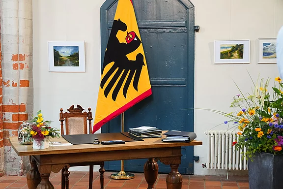 Ein hölzerner Schreibtisch steht vor einer blauen Tür und ist mit Akten und Schreibutensilien bedeckt. Hinter dem Tisch befindet sich ein Stuhl. An einer Wand hängt eine große Flagge mit dem deutschen Wappen, daneben sind einige Bilder und ein Blumenarrangement auf dem Tisch.