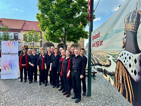 Etwa 16 Chorsängerinnen und -sänger in schwarzer Kleidung, teils mit roten Schals, posieren lächelnd für ein Gruppenfoto auf einem Platz im Freien. Ein Chor-Banner steht daneben. Szene wirkt wie vor oder nach einem Auftritt.