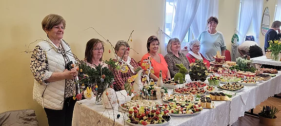 Mehrere Frauen stehen hinter einem langen Buffet voller liebevoll angerichteter Häppchen, Obstspieße, Kuchen und herzhafter Snacks. Frühlings- und Osterdekoration mit Zweigen, Moosfiguren und bunten Eiern schmückt den Tisch. Helle, freundliche Atmosphäre wie bei einer Gemeinschaftsfeier.