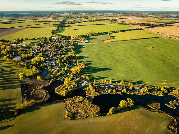 Luftaufnahme eines kleinen Dorfes, umgeben von weiten Feldern und Seen. Grüne Wiesen und Laubbäume prägen die Landschaft, während eine Straße durch das Dorf führt. Im Hintergrund sind Windkraftanlagen erkennbar, die in der Ferne stehen.