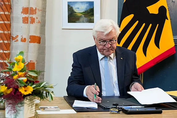 Ein älterer Mann mit grauen Haaren und Brille sitzt an einem Tisch und unterzeichnet Dokumente. Im Hintergrund hängt eine deutsche Flagge und ein Gemälde ist sichtbar. Auf dem Tisch stehen Blumen und Schreibutensilien.