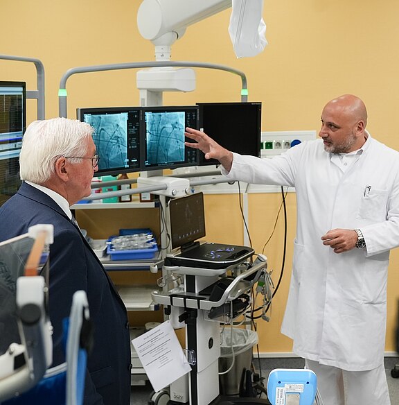 Ein Arzt in einem weißen Kittel erklärt einem älteren Mann mit grauen Haaren eine medizinische Ausrüstung in einem Behandlungsraum. Im Hintergrund sind mehrere Computerbildschirme zu sehen, die medizinische Bilder anzeigen. Der Arzt zeigt mit der Hand auf die Bildschirme.