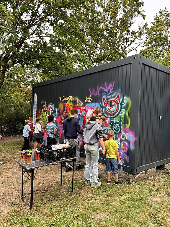 Kinder und Erwachsene besprühen gemeinsam die Wand eines grauen Containers mit bunter Graffiti-Kunst. Sie arbeiten im Freien, nutzen Spraydosen und gestalten Figuren und Schriftzüge in einer Art Workshop oder Gemeinschaftsaktion.