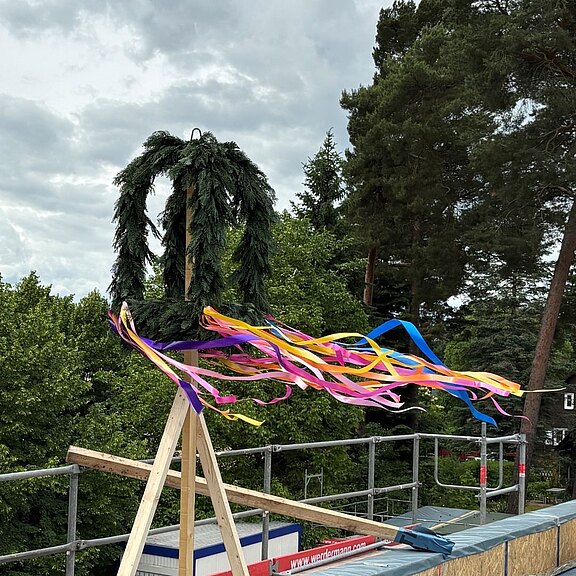 Richtkranz aus Tannengrün mit bunten Bändern auf einem Dach im Bau; die Bänder wehen im Wind.