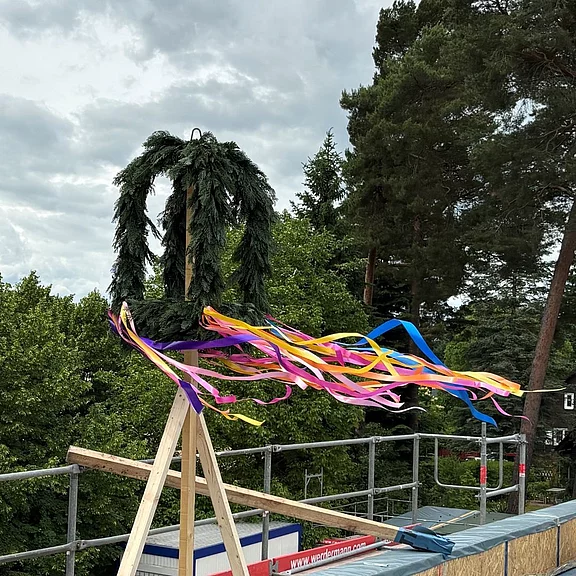 Richtkranz aus Tannengrün mit bunten Bändern auf einem Dach im Bau; die Bänder wehen im Wind.