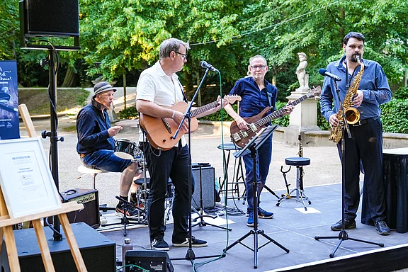 Eine Band steht auf einer Bühne im Freien und spielt Musik. Ein Mann spielt Gitarre, ein anderer Bass, ein Schlagzeuger sitzt und ein Musiker spielt Saxophon. Im Hintergrund sind Bäume und eine Statue zu sehen.