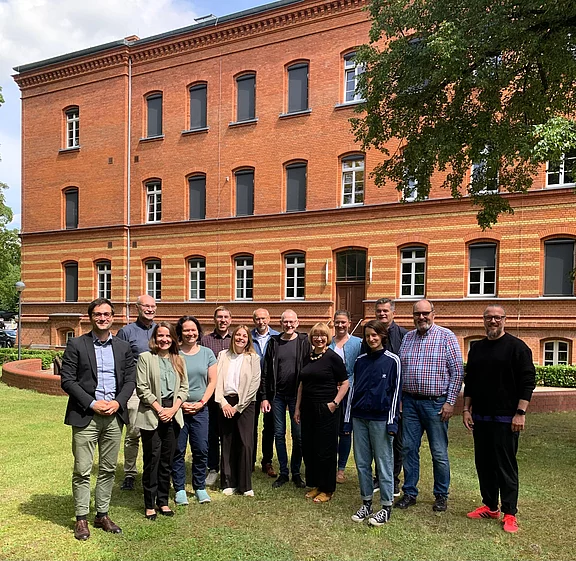 Gruppe von Erwachsenen posiert lächelnd auf einer Rasenfläche vor einem roten Backsteingebäude; informelles Gruppenfoto im Freien.