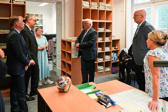 Die Abbildung zeigt eine Gruppe von fünf Personen in einem Büro. Sie stehen im Gespräch, während Aktenregale im Hintergrund sichtbar sind. Auf dem Tisch sind einige Büroartikel verteilt. Einigen Personen tragen Anzüge, während eine Person in einem hellen Kleid gekleidet ist.
