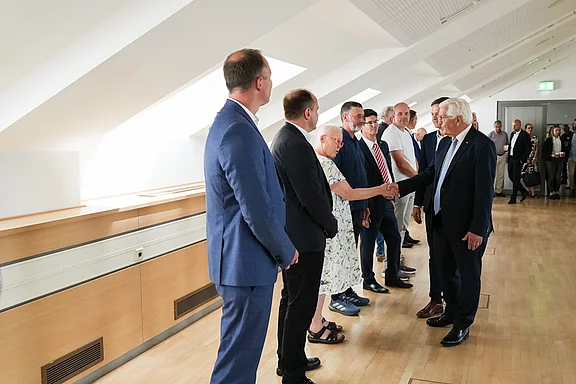 Eine Gruppe von Menschen steht in einer hellen, modernen Umgebung. Eine Person, die einen Anzug trägt, schüttelt die Hand einer anderen Person. Die Atmosphäre wirkt freundlich und förmlich, und im Hintergrund sind weitere Teilnehmer sichtbar.