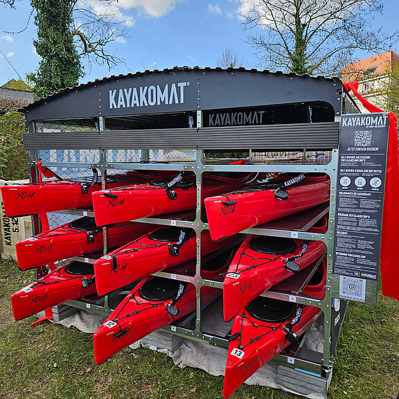 Selbstbedienungsstation „KAYAKOMAT“ mit mehreren roten Kajaks, ordentlich in Fächern eines Metallregals gestapelt. Die Boote sind startbereit und nummeriert; daneben Infotafel mit QR‑Code für Ausleihe. Outdoor-Aufstellung.