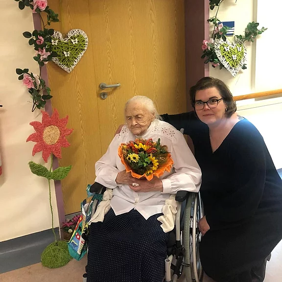 Ältere Frau im Rollstuhl hält einen Blumenstrauß; jüngere Frau hockt neben ihr und legt den Arm um sie; festlich geschmückter Flur im Hintergrund.