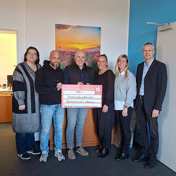 Sechs Erwachsene stehen in einem Büro und halten gemeinsam einen großen roten Spendenscheck in die Kamera – ein Gruppenfoto bei einer Spendenübergabe.