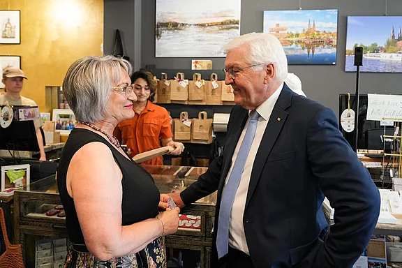Eine Frau und ein Mann stehen sich in einem Geschäft gegenüber und unterhalten sich freundlich. Im Hintergrund sind weitere Personen und einige Bilder an der Wand zu sehen, die die Umgebung des Geschäfts darstellen. Es herrscht eine freundliche, ungezwungene Atmosphäre.