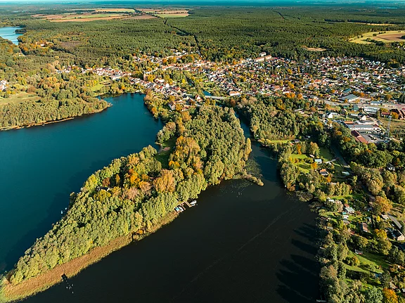 Luftaufnahme eines malerischen Dorfes, das an einem Fluss und Seen liegt. Umgeben von Wäldern und buntem Herbstlaub zeigt die Landschaft eine ruhige, natürliche Umgebung mit Wasserflächen und grünen Uferzonen. Einige Boote sind am Rand zu sehen.