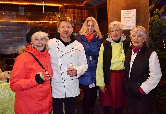 Eine Gruppe von fünf Personen steht zusammen in einem festlichen Umfeld. Sie lächeln und tragen warme Winterkleidung. Ein Mann in einer Kochjacke ist in der Mitte, umgeben von Frauen in unterschiedlichen Jacken und Mützen. Es herrscht eine fröhliche Stimmung.